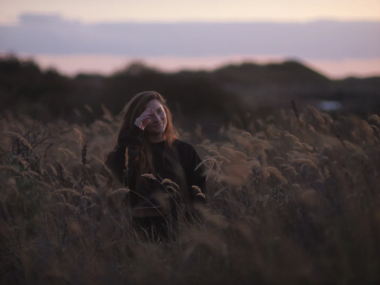Een vrouw tussen wuivende grassen, met een roze avondlucht boven de duinen